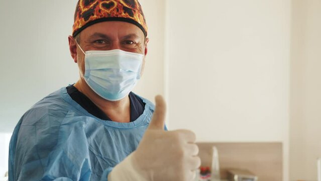 Portrait of the Professional Surgeon Looking Into Camera and Smiling after Successful Operation. In the Background Modern Hospital Operating Room. Smiling doctor showing thumbs up.