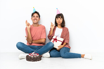 Young mixed race couple celebrating a birthday sitting on the floor isolated on white background with fingers crossing and wishing the best