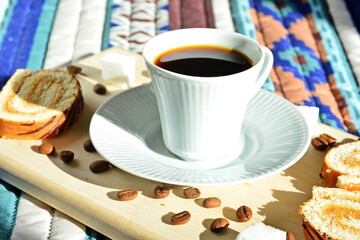 breakfast in bed - cup of strong invigorating black coffee on background of fresh sweet pastries and coffee beans with sugar cubes on wooden board on pattern cloth in bright morning sunlight close up