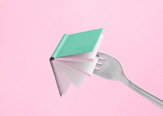 Minimal abstract composition made of fork holding a green book on isolated pastel pink background with copy space. Note card for book fair, promotions or education. Concept of reading or learning. 