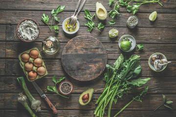 Food background with healthy ingredients: spinach bunch, avocado, spices, lime, eggs,  and kitchen utensils, cutlery and empty round cutting board on rustic wooden kitchen table. Top view.