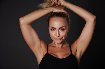Sexy middle aged woman with perfect body, beautiful healthy tanned skin, posing in black lingerie and tying a ponytail on her head, three quarter studio portrait over dark background with copy space