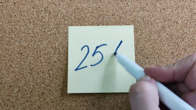 Handwritten inscription "25%" in blue felt-tip pen on a yellow paper sticker. Sticker on a cork board close-up. Blue marker in a woman's hand