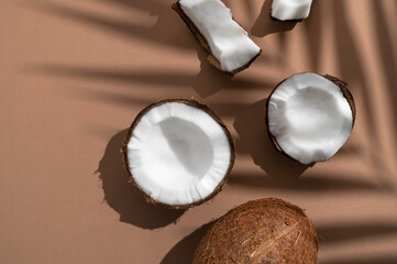 Fresh whole and broken pieces tropical coconut nut with sunny contrast shadows palm leaf on brown background flat lay top view. Creative summer food background, exotic organic healthy diet fruit