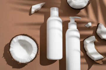 White cosmetic bottles broken fresh coconut with sunny contrast shadows palm leaf on brown background. Organic natural cosmetics with coconut oil. Plastic packaging products for branding, mock up