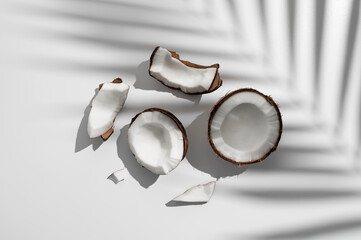 Fresh whole and broken pieces tropical coconut nut with sunny contrast shadows palm leaf on light gray background flat lay top view. Creative summer food background, exotic organic healthy diet fruit