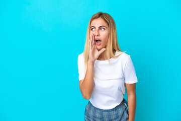 Blonde Uruguayan girl isolated on blue background whispering something with surprise gesture while looking to the side