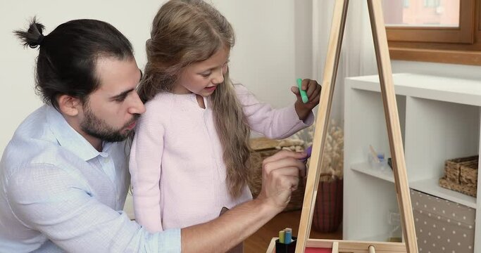 Loving father and little daughter holding colored chalks drawing together on board, close up. Young dad and preschool child having fun at home, involved in creative hobby. Art classes, daycare concept