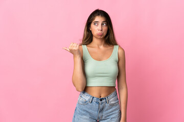 Young caucasian woman isolated on pink background unhappy and pointing to the side