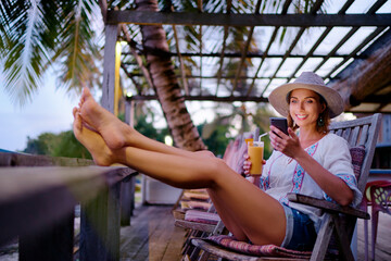 Vacation and technology. Young pretty woman in hat using smartphone sitting at beach cafe bar.