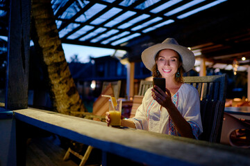 Vacation and technology. Young pretty woman in hat using smartphone sitting at beach cafe bar.