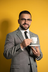 Handsome bearded young man in eye glasses in formal wear holding a cup of coffee isolated on yellow background. Business, hot drinks and people concept