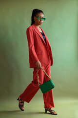 Fashionable Black woman wearing trendy orange suit, green sunglasses, holding stylish leather bag posing on green background. Full-length fashion studio portrait, natural light - 496622432