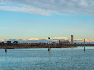 View from Burano across the water towards the island of Torcello and the church of Basilica di Santa Maria Assunta with the snow-capped Dolomite mountains in the background - Torcello, Venice, Italy