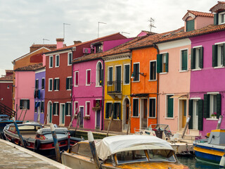 A row of brightly colored houses face the canal on the beautiful Venetian island of Burano, boats...