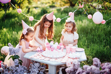 Summer outdoor kids birthday party. Group of happy Children celebrating birthday in park. Children blow candles on birthday cake. Kids party pink pastel decoration and food. Presents and sweets.