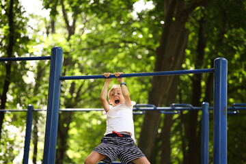 Fototapeta na wymiar the boy goes in for sports in the park hangs on the horizontal bar pulls himself up and shouts loudly