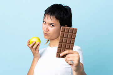 Young Uruguayan woman over isolated blue background having doubts while taking a chocolate tablet...