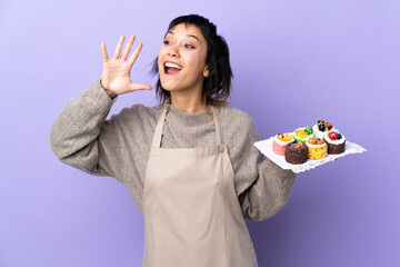 Young Uruguayan woman holding lots of different mini cakes over isolated purple background shouting with mouth wide open