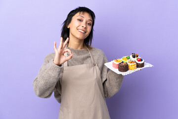 Young Uruguayan woman holding lots of different mini cakes over isolated purple background showing an ok sign with fingers