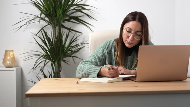 Young entrepreneur woman with eyeglasses working on laptop and writing notes on notebook in a cozy office
