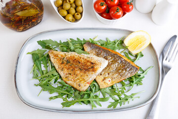 Fried dorado fillet with crispy skin. Arugula, cherry tomatoes, olives, lemon and olive oil. Traditional Mediterranean dish. White background. Selective focus, close-up, top view.