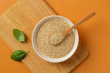 White quinoa grains and spoon in a beige bowl on a wooden cutting board over orange background. Chenopodium quinoa seeds for gluten free dieting. Concepts of superfood vegetarian healthy eating.