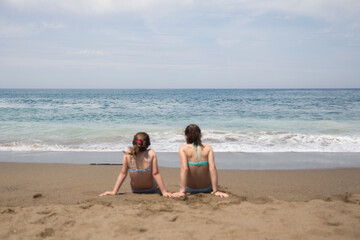 Two women sitting on the beach in swimsuits looking at the sea with a turquoise blue sky © FOTO GATO.