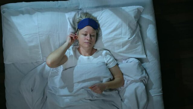 Woman before going to sleep lying in bed plugs her ears with earplugs and puts mask on her eyes, top view.