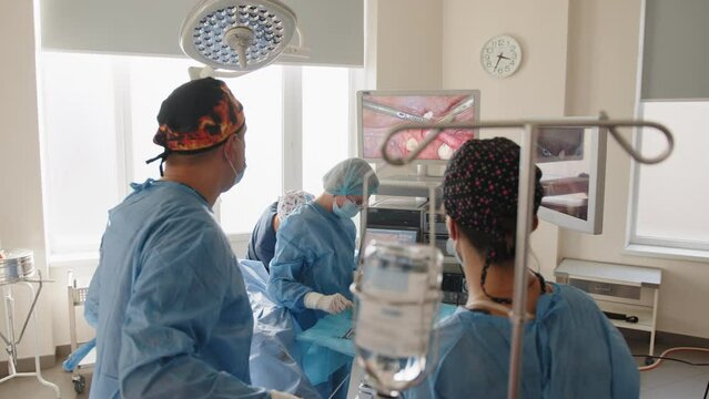 Back view of surgeons team looks at monitors while preforming operation in hospital operating theater, male surgeon operating patient working with surgical laparoscopy instruments. Gynecology