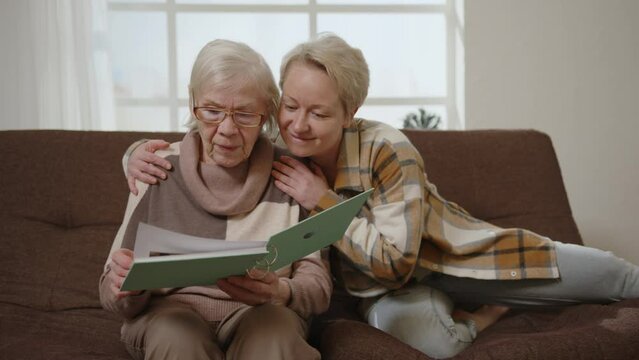 Loving daughter with elderly mother look at photo album in an embrace, enjoying the memories. Two generations of adult women.
