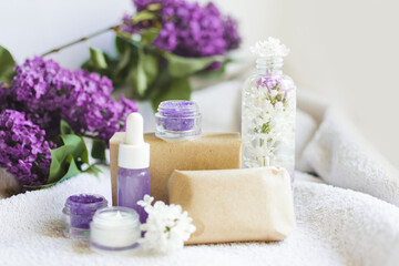Soup in craft paper, aroma oil, sugar scrub, cream with lilac flowers on white terry towel background. Mockup and copy space. Spa treatments for home care. Home rejuvenation and moisturizing