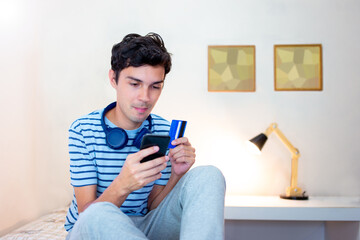 Man sitting on bed at home and using mobile app for online shopping and using credit card to pay. Online food delivery. On the wall, digitally created mosaics to simulate two paintings.