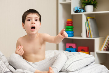 A 4-year-old boy with his mouth open sits on the bed and points his finger to the side against the background of a cabinet with toys.