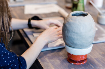 A girl is sculpting details for a clay vase, which stands nearby.