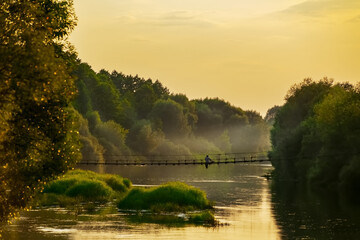 Wooden suspension bridge over a quiet river at sunset. The figure of a man is sitting on a suspension bridge over the river. The concept of loneliness