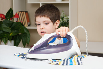 A boy of 4 years learns to hold an iron, iron clothes on an ironing board. Helper, help children to...