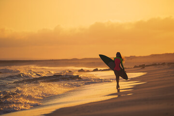Pensive young woman in swimsuit walking on the beach with surfboard in sunset. (Golden hour).