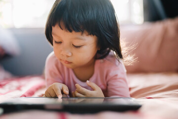 A little girl in a pink shirt lay intently playing the tablet on her bed. self-learning technology...