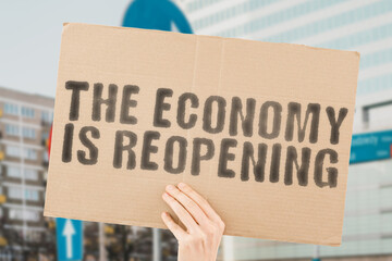 The phrase " The economy is reopening " on a banner in men's hands with blurred background. Improve. Grow. Opening. Ownership. Network. Lawyer. Panic. Political. Solution. Corrupt. End. Crisis. Office