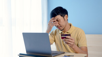 Handsome Asian man, wearing a yellow shirt sitting on a white bed, has a serious look on his face when he chooses a credit card to pay for an order online, on his laptop computer.