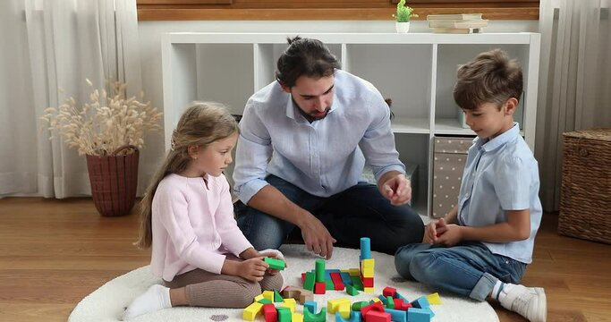 Father play wooden bricks with preschool son and daughter. Little siblings sit on floor in nursery, enjoy playtime and talk with caring young dad at home. Happy fatherhood, upbringing, leisure concept