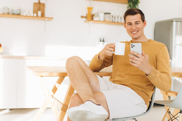 Handsome guy in yellow casual clothes sitting at home in the kitchen drinking coffee and working on tablet and phone