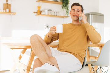 Handsome guy in yellow casual clothes sitting at home in the kitchen drinking coffee and working on tablet and phone