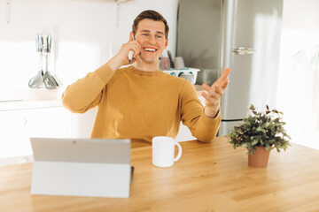 Handsome guy in yellow casual clothes sitting at home in the kitchen drinking coffee and working on tablet and phone