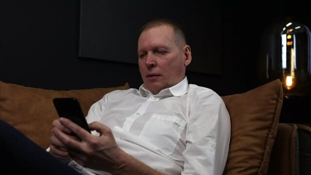 Mature businessman on sofa with mobile phone in hands, texting, reading and calling smb