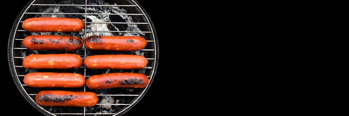 Grilling sausages on barbecue grill on black background with copy space. Sausages sizzling over the coals on grill. Top view. Suitable for creating web banners.