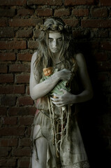 A girl is dressed in rags as a Halloween horror 
figure. She stands in front of an old brick wall. 
Her clothes are torn and dirty and she holds a 
broken doll in her arms.
