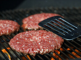 Fresh raw beef burgers on grill grate with flames and stainless steel spatula in background