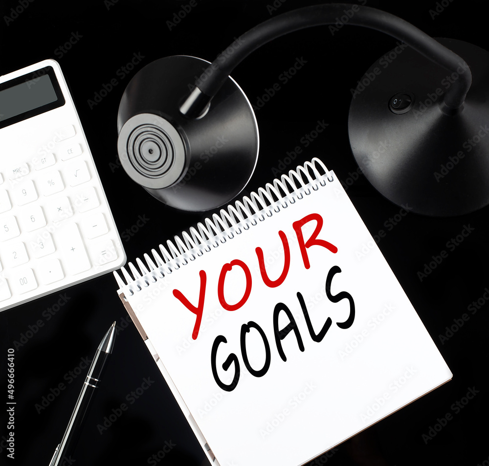 Poster opened paper notebook with text your goals with a pen, calculator and table lamp on black background